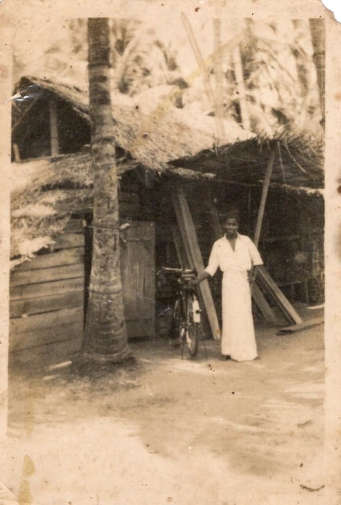 A man in a white shirt and sarong standing with a bicycle outside a palm-thatched timber shed, sawn boards leaning against the wall.