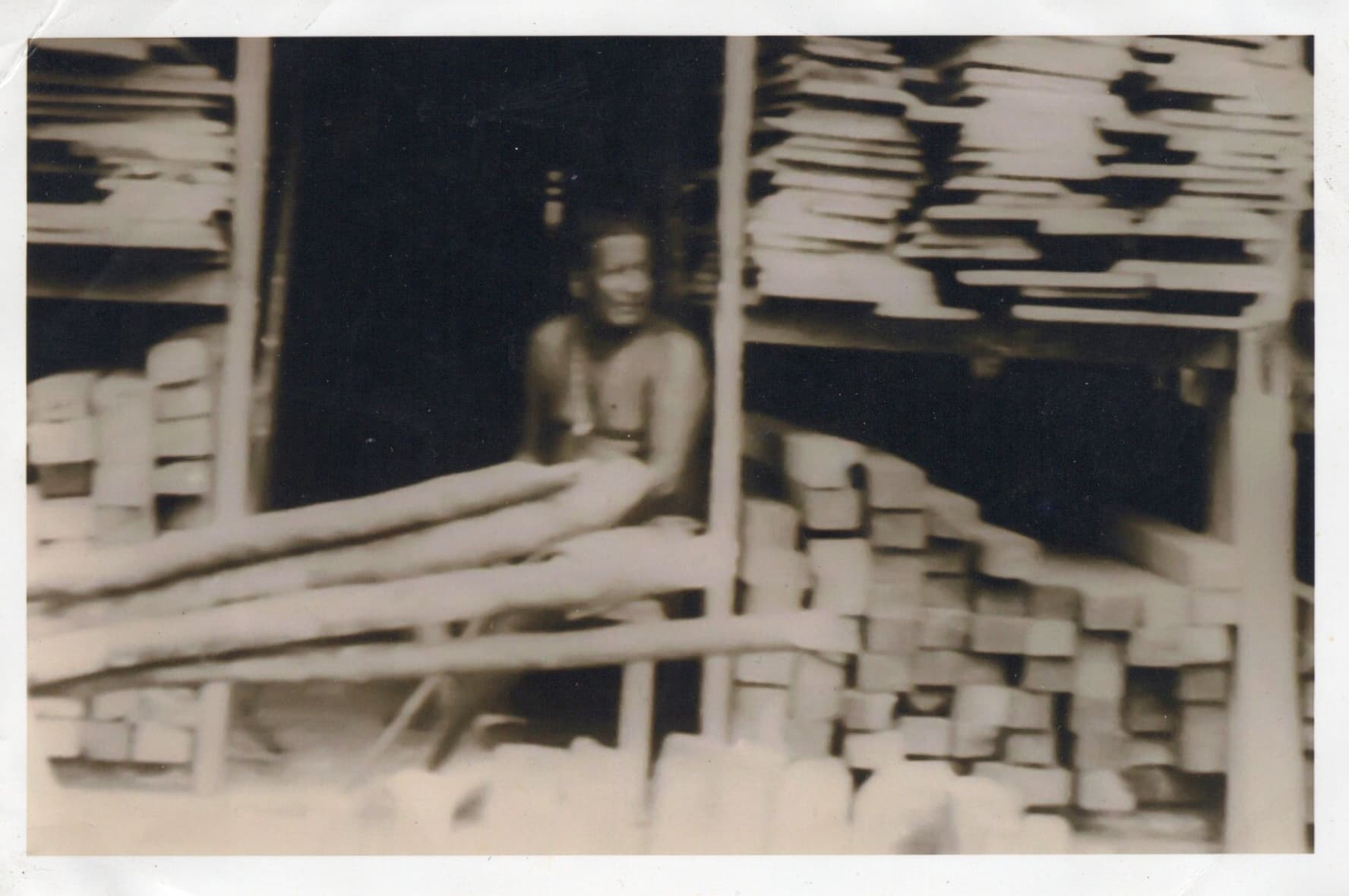 A man seated at a workbench inside the timber store, surrounded by stacked sawn timber on every side.