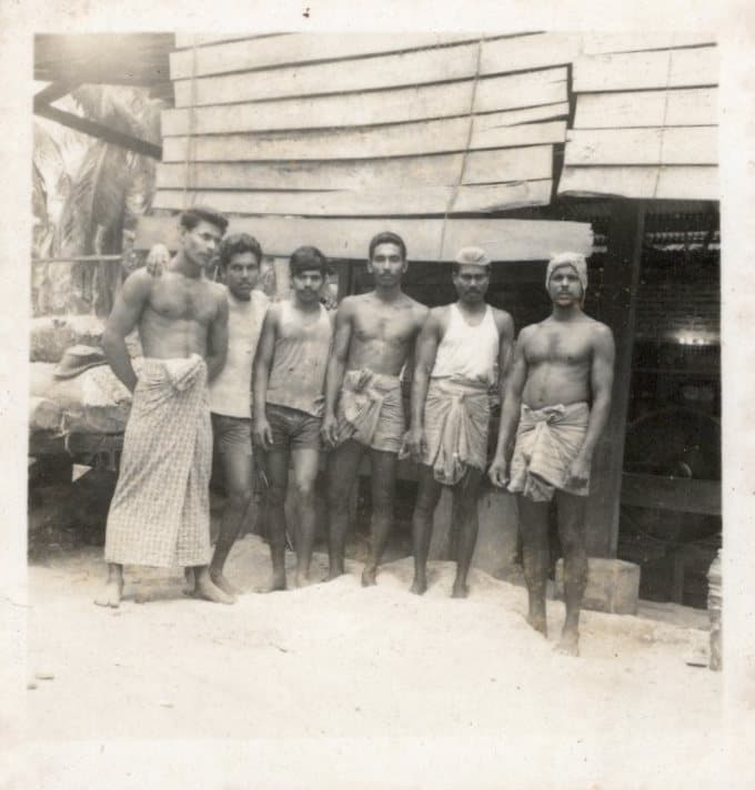 Six members of the yard team standing shoulder to shoulder outside the mill, beneath a stack of sawn boards.